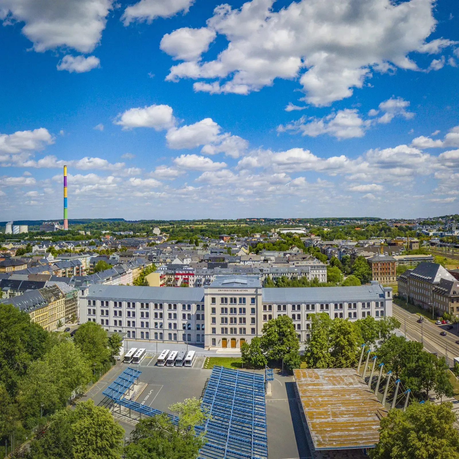 Auf dem Bild ist ein viertöckiges Gebäude im Bauhausstil, mit zwei Flügeln und einem 5 stöockend Hauptteil abgebildet, davor sind Gitte und Dächer zu erkennen, die eine Fahrradparkplatz überdecken. Rechts und Links sind Bäume zu erkennen. Im Hintergrund erkannbar ist eine grüne Stadt. Links hinten im Bild ist ein Wahrzeichen Chemnitz, ein bunt bemalter Schornstein zu erkennen. Die obere Hälfte des Bildes nimmt der sehr blaue Himmel ein mit Quellwolken. Das Bild ist im Sonnenschein fotografiert und leuchtet sehr.