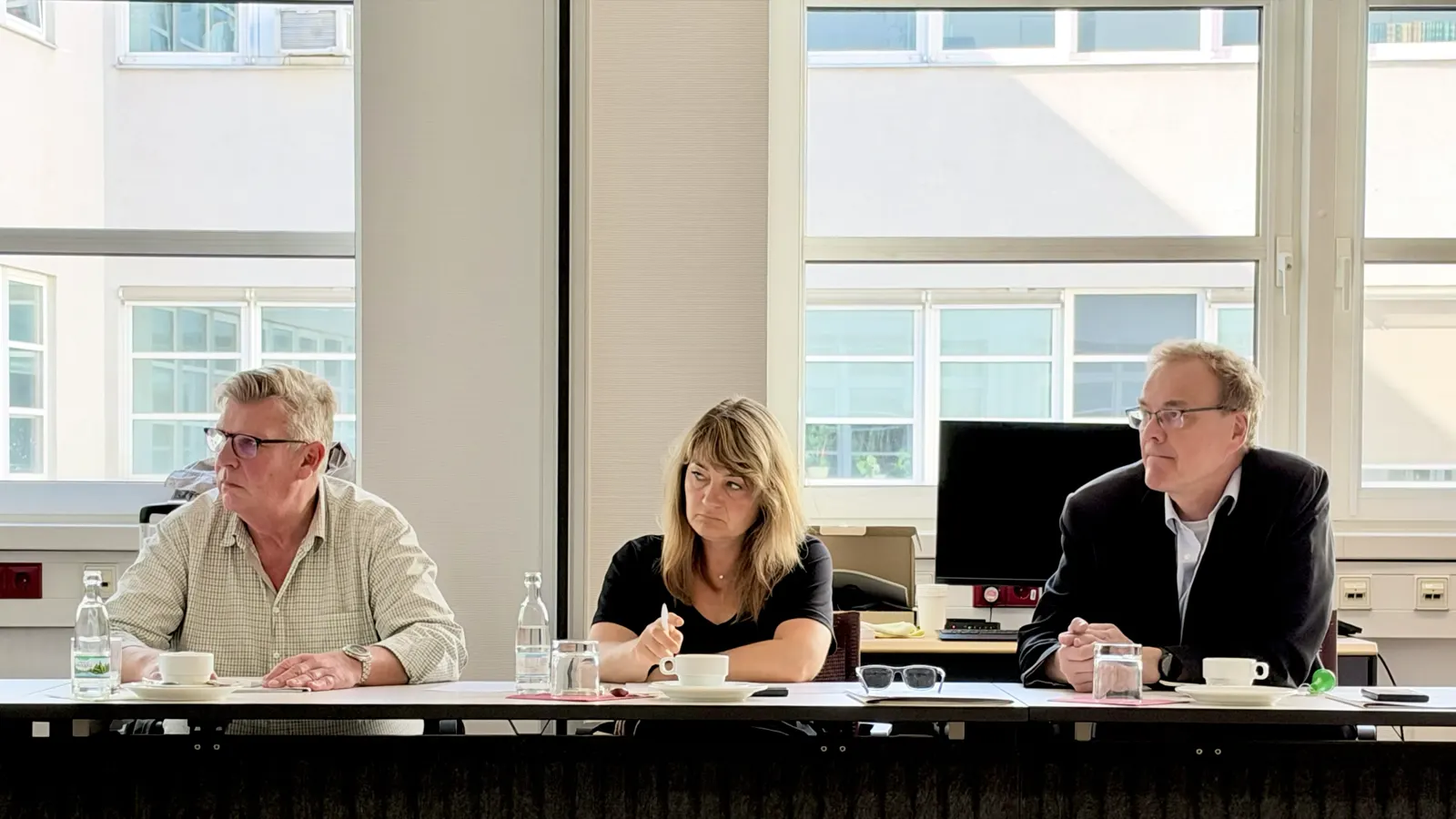 Drei Teilnehmende an einem Konferenztisch vor einer Fensterfront