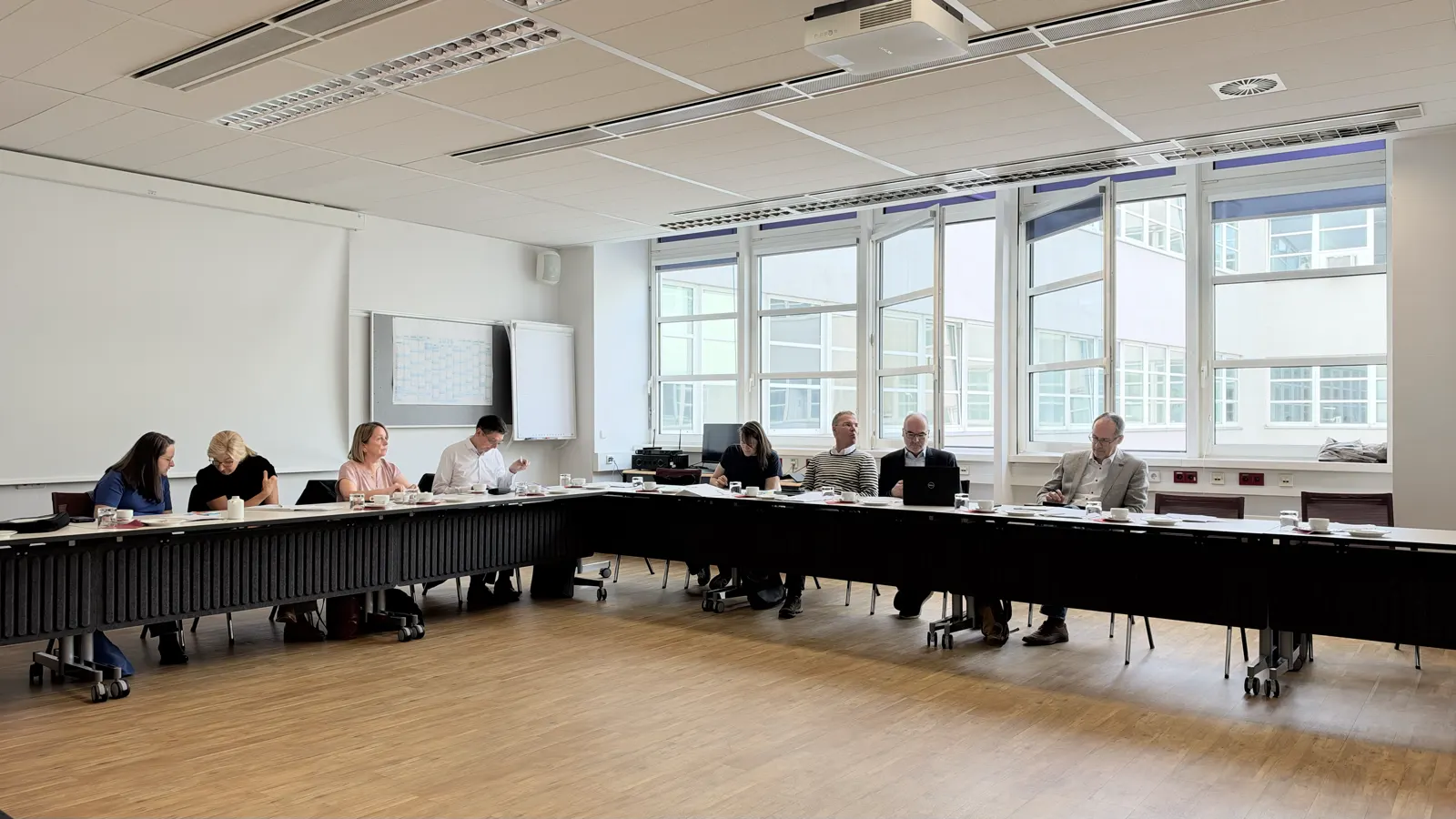 Sitzungsraum mit einer langen Tischreihe, an der Teilnehmende sitzen